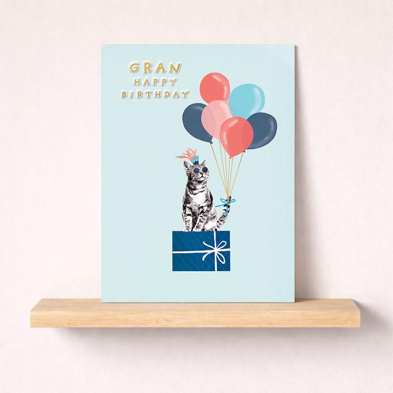 Birthday card for Gran featuring a cat wearing sunglasses and a party hat holding balloons on a pastel background