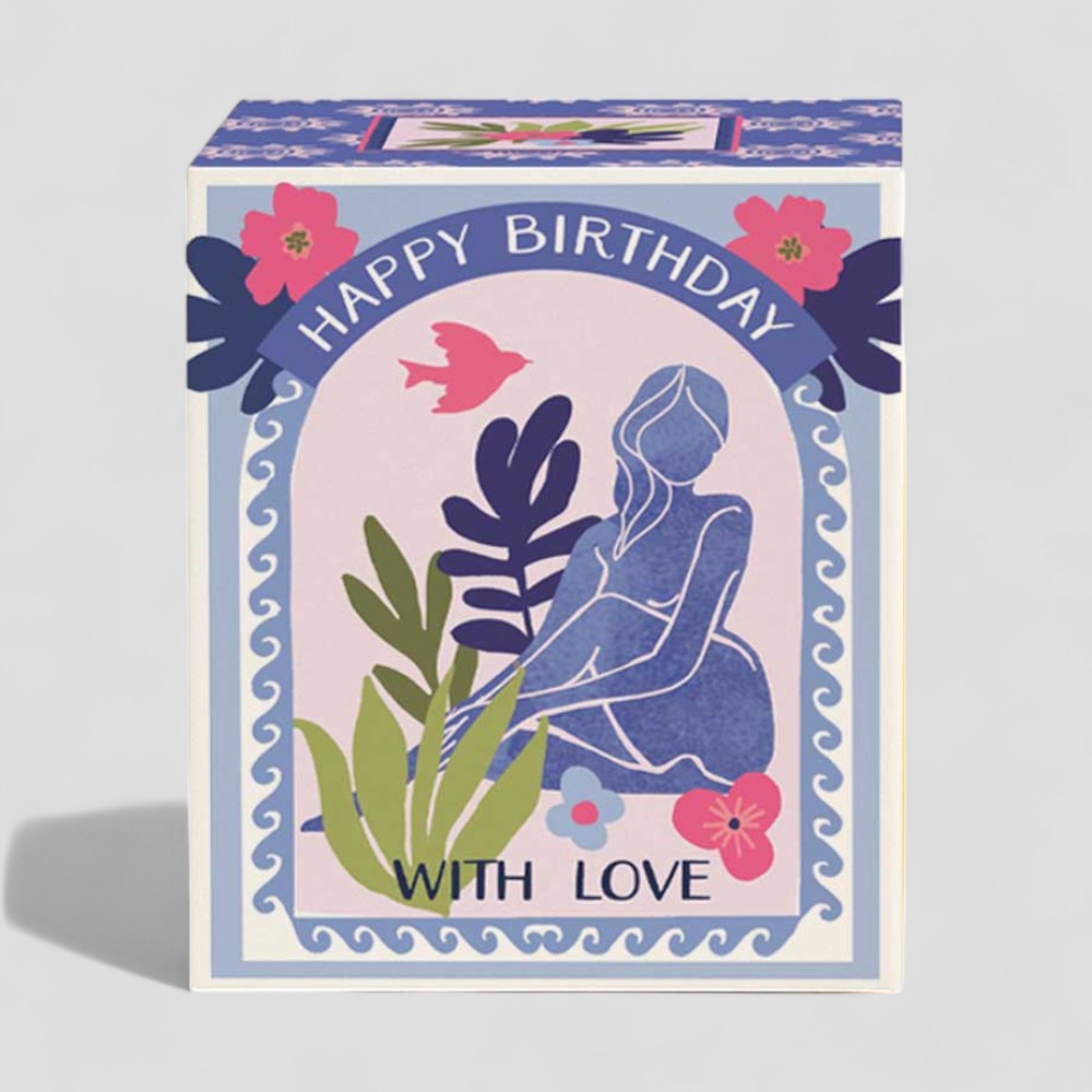 Lit yellow candle in glass beside Happy Birthday gift box with floral design