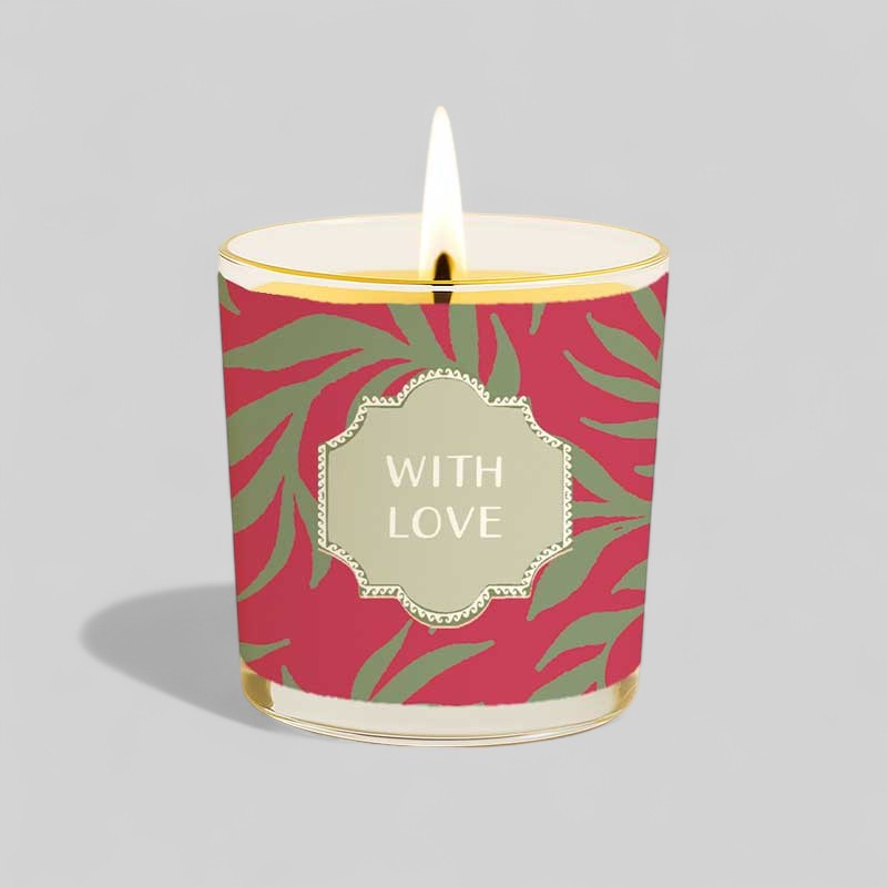 Lit yellow candle in glass with red and green patterned box saying With Love