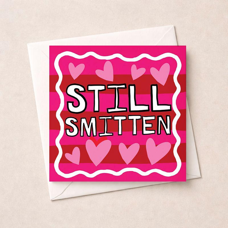 A charming greetings card with pink and red stripes, hearts, and "STILL SMITTEN". Perfect for expressing ongoing love and affection.