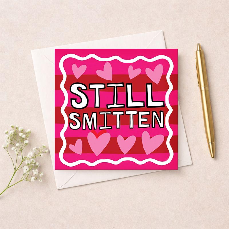 A charming greetings card with pink and red stripes, hearts, and "STILL SMITTEN". Perfect for expressing ongoing love and affection.