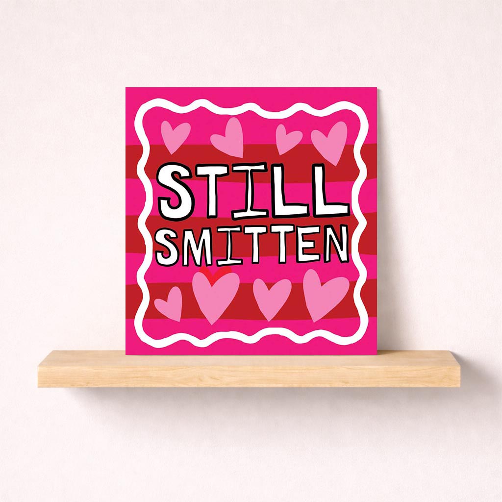 A charming greetings card with pink and red stripes, hearts, and "STILL SMITTEN". Perfect for expressing ongoing love and affection.